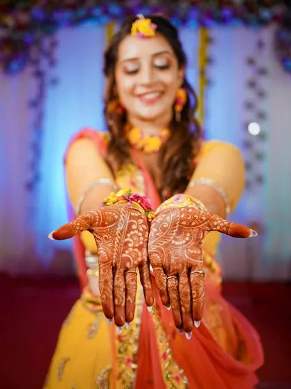 A classic Haldi shot where the bride joyfully presents her beautifully decorated mehndi hands. The focus is sharp on the intricate henna design, with the bride's happy, blurred smile in the background.