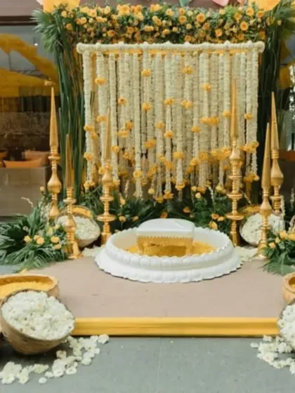 This is an elegant and traditional Haldi ceremony setup. It features a central white seat for the rituals, surrounded by bowls of fresh flower petals, all set against a beautiful backdrop of white and yellow flower strings.