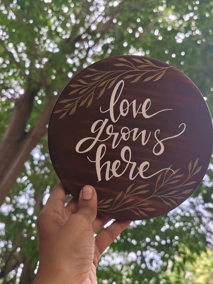 Holding our "love grows here" plaque up to the sky. The dark wood and white calligraphy stand out beautifully against the natural backdrop, a reminder of love's organic beauty.