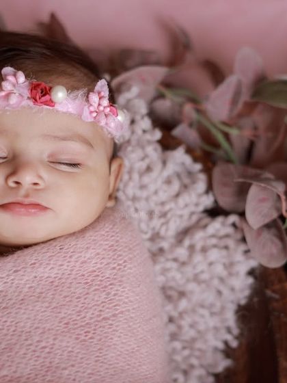 A close-up of a baby girl in a pink swaddle and floral crown, nestled in a bowl surrounded by eucalyptus and pink peonies.