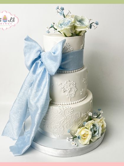 A timeless wedding cake with a touch of "something blue." This three-tiered creation features intricate white lace piping and is wrapped in a large, flowing blue fabric bow.