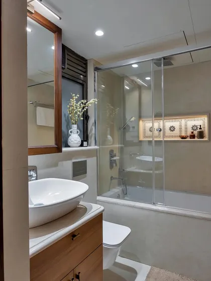 This cozy bathroom features a bathtub with a glass enclosure, a wooden vanity, and warm lighting. A small niche with decorative objects adds a personal touch.
