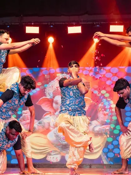 A traditional dance performance with a human pyramid formation. The stage is lit with warm spotlights, and a graphic of Lord Ganesha is displayed on the LED screen behind them.