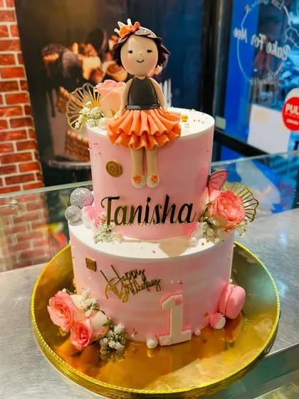 A two-tier pink first birthday cake with a beautiful doll topper and fresh floral decorations.
