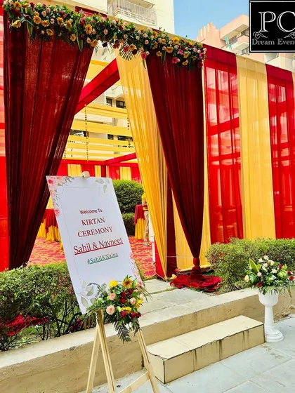 The entrance to a Kirtan ceremony, decorated in traditional red and yellow drapes and florals. A personalized welcome sign on an easel greets guests as they arrive for the sacred event.