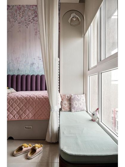 A cozy window seat provides the perfect spot for reading. The bed features a deep purple fluted headboard set against a floral wallpaper, and sheer curtains can be drawn to create a private nook.