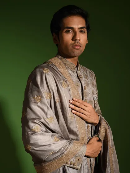 My steel grey raw silk sherwani offers a modern take on groomswear, detailed with ivory thread, bronze zari, and delicate stonework. The matching stole completes a look that is both understated and grand.