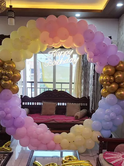 A beautiful pastel-colored circular balloon arch set up in a living room. The neon 'Happy Birthday' sign in the center adds a nice touch.