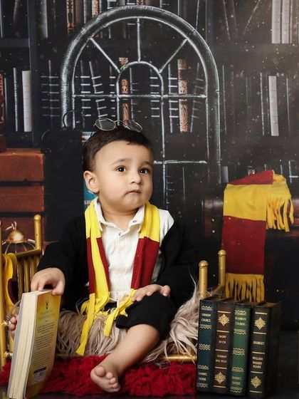 A moment of quiet contemplation for our little wizard. This pose shows off the full Gryffindor-style costume against the rich, dark library backdrop, perfect for a themed baby portrait.
