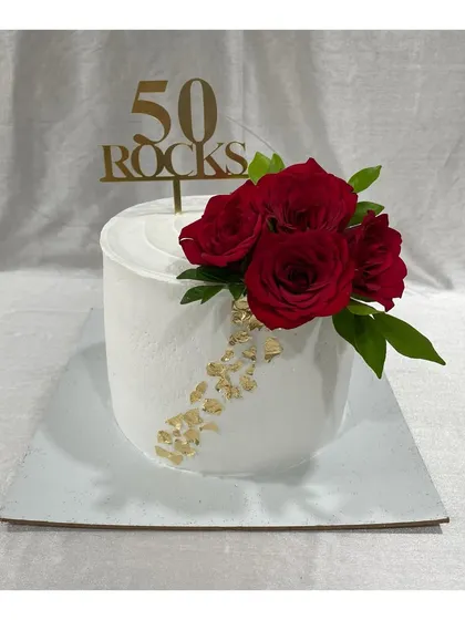 A classy cake for a 50th birthday with the message "50 Rocks". The clean white finish is accented with gold leaf and a bouquet of fresh red roses.