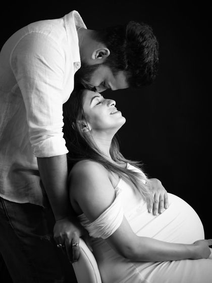 An intimate black and white portrait. The partner kisses the mom-to-be's forehead as she sits, both sharing a peaceful and loving moment.