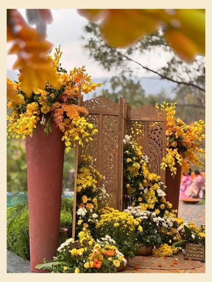 An earthy and elegant haldi backdrop. Set against a mountain view, this decor features a carved wooden screen and large terracotta pots overflowing with yellow and orange marigolds.
