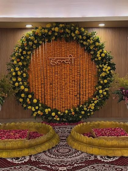 An indoor Haldi setup with a large marigold ring and a neon "Haldi" sign. The two lotus-shaped urlis are filled with rose petals, creating an elegant and modern look.