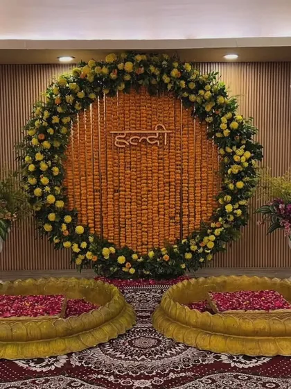 An indoor Haldi setup with a large marigold ring and a neon "Haldi" sign. The two lotus-shaped urlis are filled with rose petals, creating an elegant and modern look.