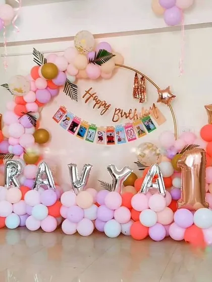 A charming and personalized first birthday decoration for Ravya. This home setup features a pastel-colored balloon arch on a gold ring stand, with the baby's name in silver foil letters and a monthly milestone photo banner.