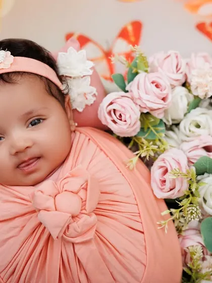 A lovely newborn girl, swaddled in pink, looks at the camera from a bed of flowers. Her alert expression is just priceless.