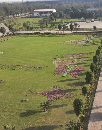 An aerial view of the massive lawn at the Jhajjar Power site, featuring large-scale floral art, including butterfly shapes and the words "SAFETY 1st", created with thousands of flowers.