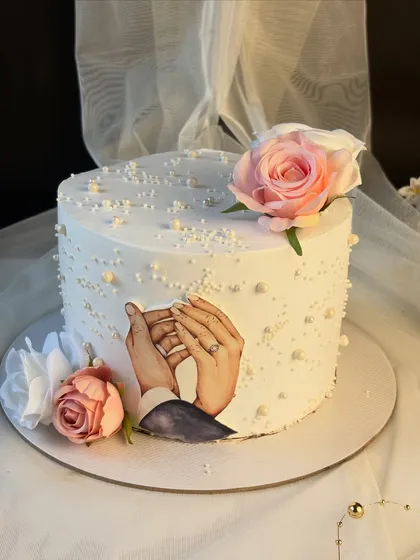 This elegant single-tier cake was created to celebrate a 25th wedding anniversary. It features a beautiful edible print of the couple holding hands, surrounded by delicate sugar pearls and soft pink and white silk flowers.