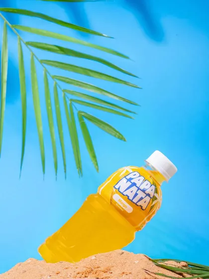 A bright, summery shot of a Papa Nata lemon drink on a mini beach setup. The vibrant blue and yellow colors make the product pop, perfect for a refreshing beverage brand.