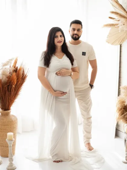 A stylish and serene portrait of an expecting couple. Dressed in coordinating white outfits, they stand together in a bohemian-themed studio, radiating calm and happiness.