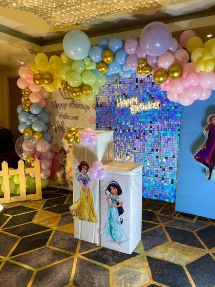 A Disney Princess themed birthday party featuring a blue iridescent shimmer wall. The decoration is completed with a pastel balloon arch, a 'Happy Birthday' neon sign, and pedestals with images of Princess Jasmine and Snow White.