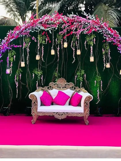 This outdoor stage setup is bursting with color. A deep green hedge wall provides the perfect canvas for a vibrant arch of pink and purple cherry blossoms and hanging lights.