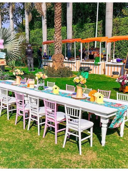 Long dining tables set for a fairy feast in the garden. The setup includes themed table runners and views of the various activity and food stalls in the background.