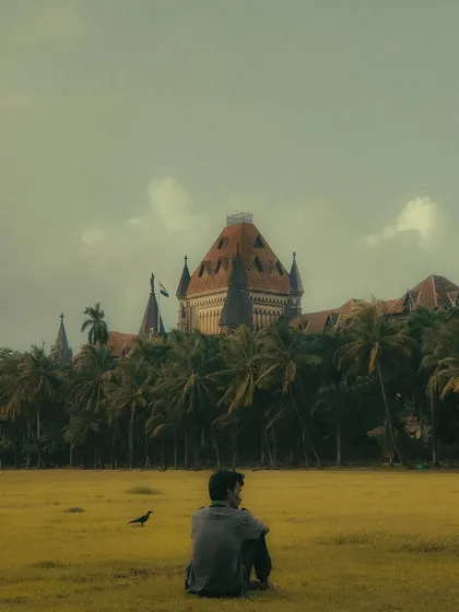 A young man sits alone in the vast green expanse of Oval Maidan, with the Bombay High Court in the background. This image captures a powerful sense of solitude and introspection within a public space.