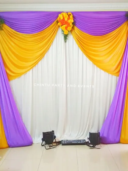 A simple drape decoration for an engagement at home. The backdrop uses purple and yellow fabric with small floral arrangements.