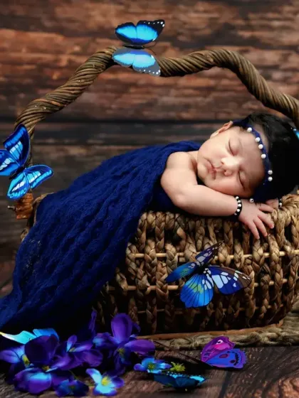 A newborn sleeps peacefully in a rustic basket, wrapped in a deep blue cloth and surrounded by butterfly props.
