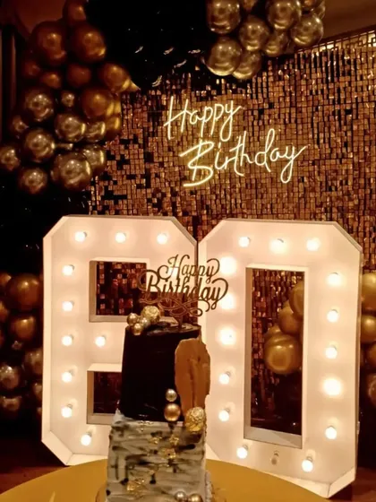 For a glamorous 60th birthday, I created this stunning photo corner. It features a gold sequin shimmer wall, a custom 'Happy Birthday' neon sign, and large marquee light-up numbers, all framed by a rich black and gold balloon garland.