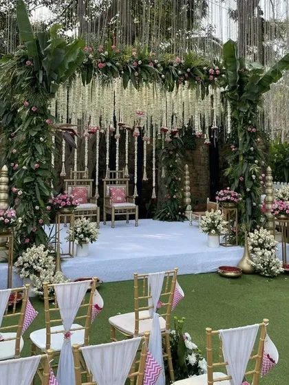 A beautiful and traditional mandap design for an outdoor ceremony. The structure is adorned with lush banana leaves, pink flowers, and hanging tuberose strings, creating a fresh and authentic South Indian wedding vibe.