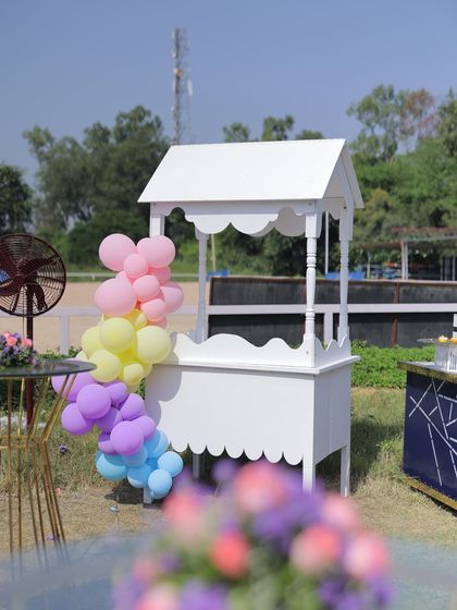 A classic white cart decorated with a pastel balloon garland, ready to be used as a drink or dessert station at an outdoor carnival party.