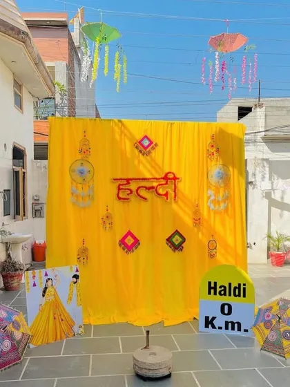 An outdoor Haldi photo corner complete with fun and quirky props. The setup includes a bright yellow backdrop, hanging Rajasthani umbrellas, and a custom "Haldi 0 Km" milestone sign for playful pictures.