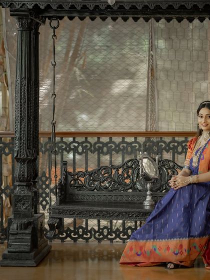 This model showcases a beautiful Paithani silk saree in blue and orange, seated on a traditional Indian swing.