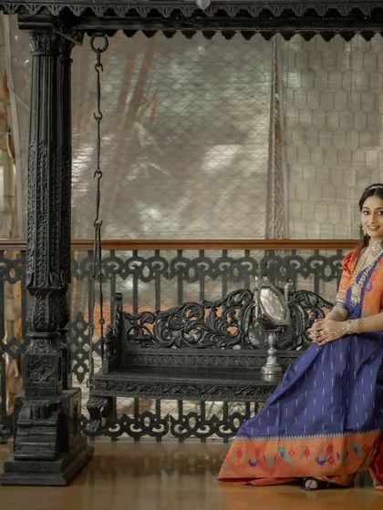 This model showcases a beautiful Paithani silk saree in blue and orange, seated on a traditional Indian swing.