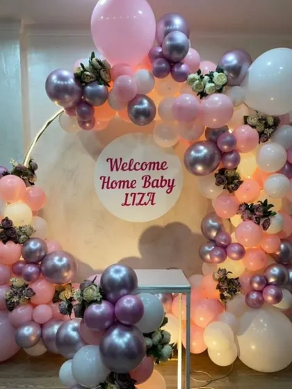 A personalized "Welcome Home Baby" ring decoration in pastel pink, white, and metallic purple. The custom name in the center makes it extra special.