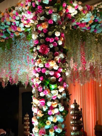 A close-up view of a mandap pillar completely covered in a dense arrangement of pink, peach, and white flowers. This technique creates a lush, textured look and feel, complemented by hanging white floral strings.