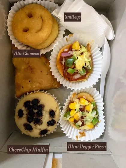 A close-up of a birthday party snack box with a choco-chip muffin, mini veggie pizza, mini samosa, and smileys.
