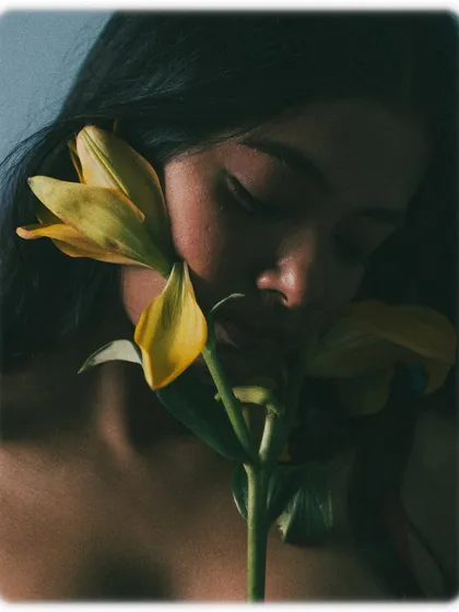 A quiet, contemplative moment. The model's eyes are closed, and her face is gently turned towards the flower. The deep shadows and soft highlights create a moody, painterly effect.