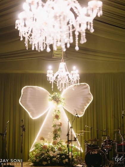 A close-up of the illuminated bow on stage, adorned with florals and set against a backdrop of sparkling chandeliers, showcasing the central design element of the "Big Bow Affair."