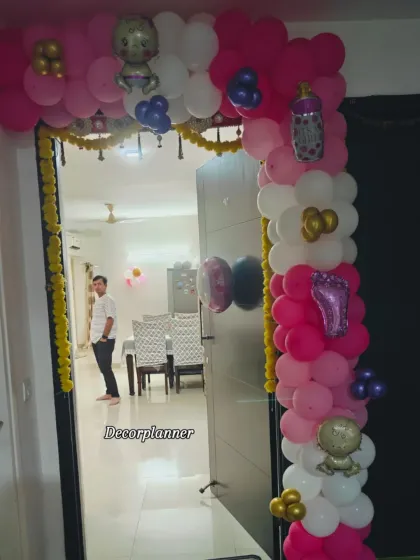 The entrance to a home decorated for a baby girl's welcome. The pink, white, and gold balloon arch is accented with baby-themed foil balloons and traditional marigold garlands for a festive touch.