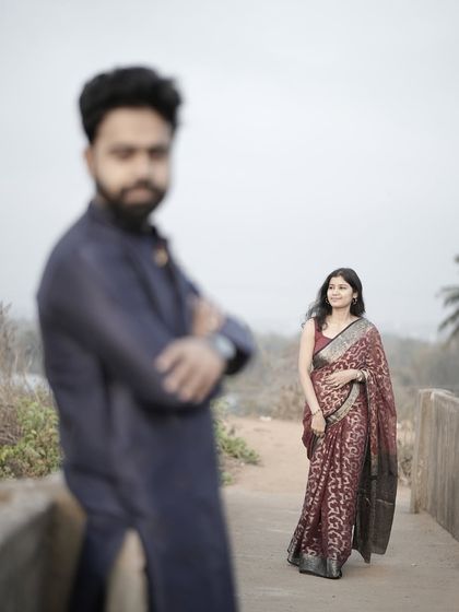 An artistic shot with the groom out of focus in the foreground, drawing the eye to the bride in her beautiful red saree.