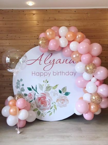 A floral-themed birthday backdrop with a personalized print and a delicate pink and white balloon arch.