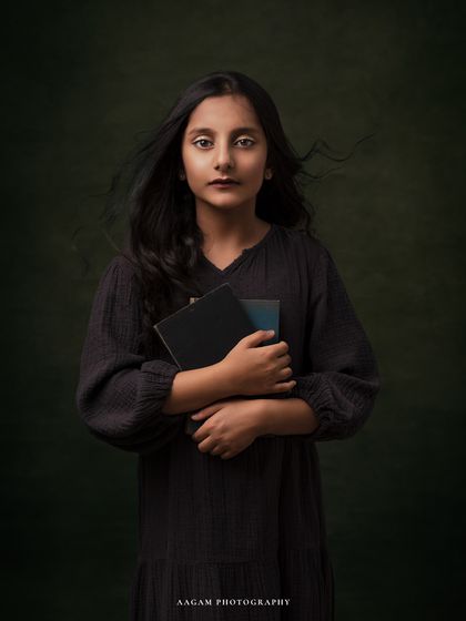 A moody and emotive fine art portrait of a young girl holding books. The wind in her hair and the dark, textured backdrop create a feeling of story and introspection.
