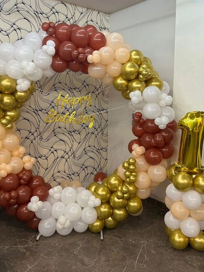 This first birthday decoration features a large balloon ring in neutral and metallic tones. The arrangement includes white, beige, brown, and gold chrome balloons, with a number '1' foil balloon column on the side.