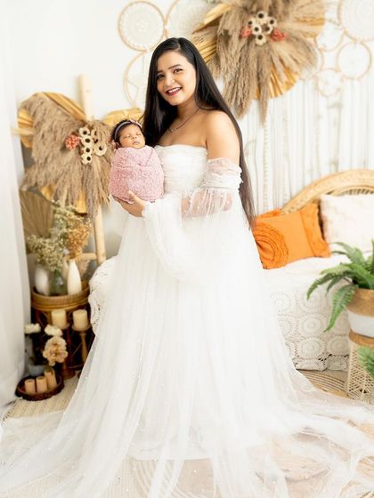 A radiant mother holds her newborn daughter. Dressed in a flowing white gown, she stands in our bright, boho-chic studio, creating a portrait that is both angelic and warm.