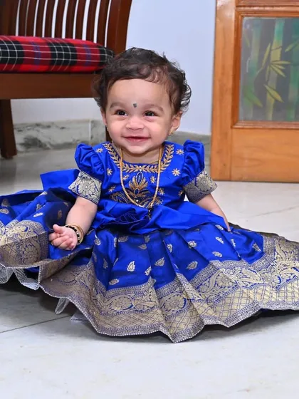 A smiling little one in her bespoke Pattu Pavadai. This royal blue silk dress, adorned with traditional gold zari work and puffed sleeves, is designed for both beauty and comfort, making it ideal for cultural ceremonies, festivals, and first birthdays.