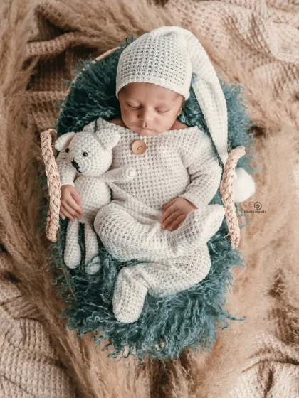 A sleeping newborn in a cozy, textured white romper and matching hat, holding a small knitted toy. This boho-inspired shot uses natural textures like pampas grass for a warm, earthy feel.
