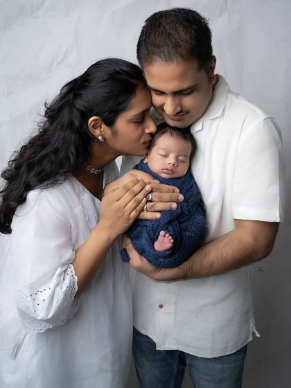 A mother's gentle kiss and a father's protective embrace. This intimate family portrait, with the baby swaddled in a cozy blue wrap, is full of warmth and emotion.